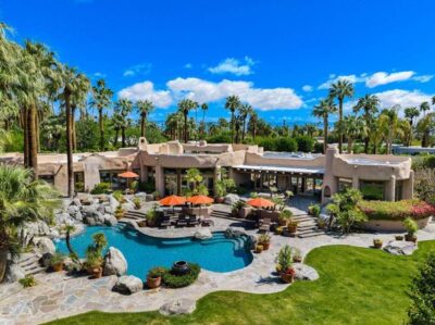 Luxury desert estate with lagoon-style pool, rock landscaping, palm trees, and mountain views under a bright blue sky