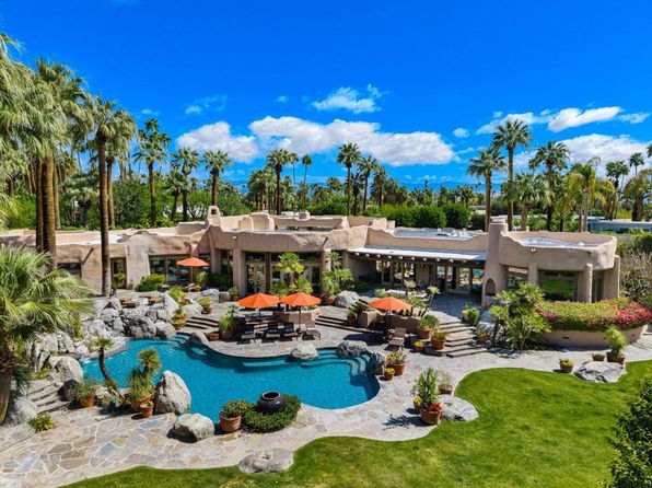 Luxury desert estate with lagoon-style pool, rock landscaping, palm trees, and mountain views under a bright blue sky