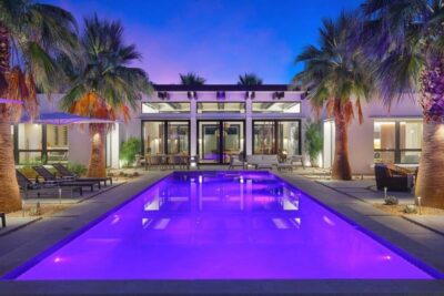 Modern Palm Springs desert home at dusk with a long rectangular swimming pool glowing purple, framed by tall palm trees.