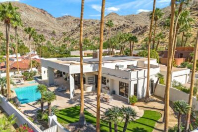 Modern Palm Springs–style desert home with a turquoise swimming pool, tall palm trees, and rugged mountain backdrop under a blue sky.